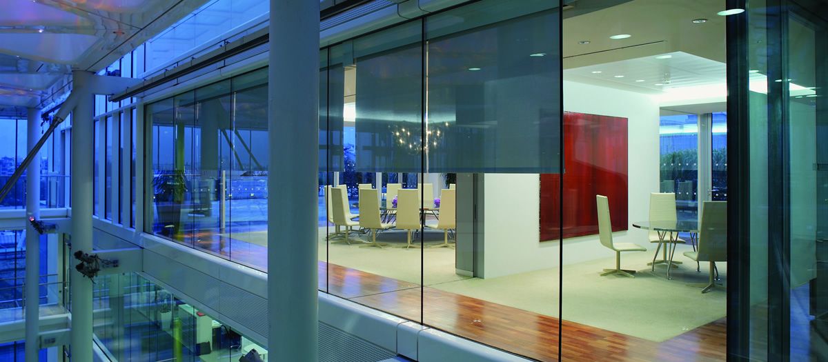 Glass-walled conference room in a modern office with roller shades, contemporary chairs, and city views at dusk.