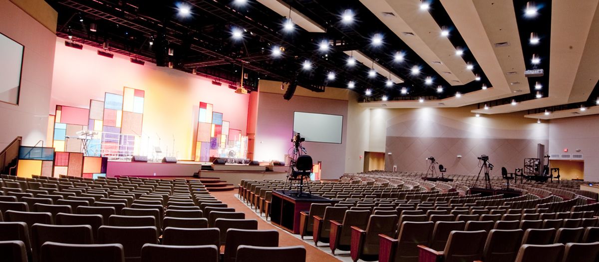 Large auditorium with rows of empty seats facing a brightly lit stage framed by colorful geometric panels; ceiling packed with spotlights and suspended speakers, with several video cameras on tripods positioned along the center aisle for live production.