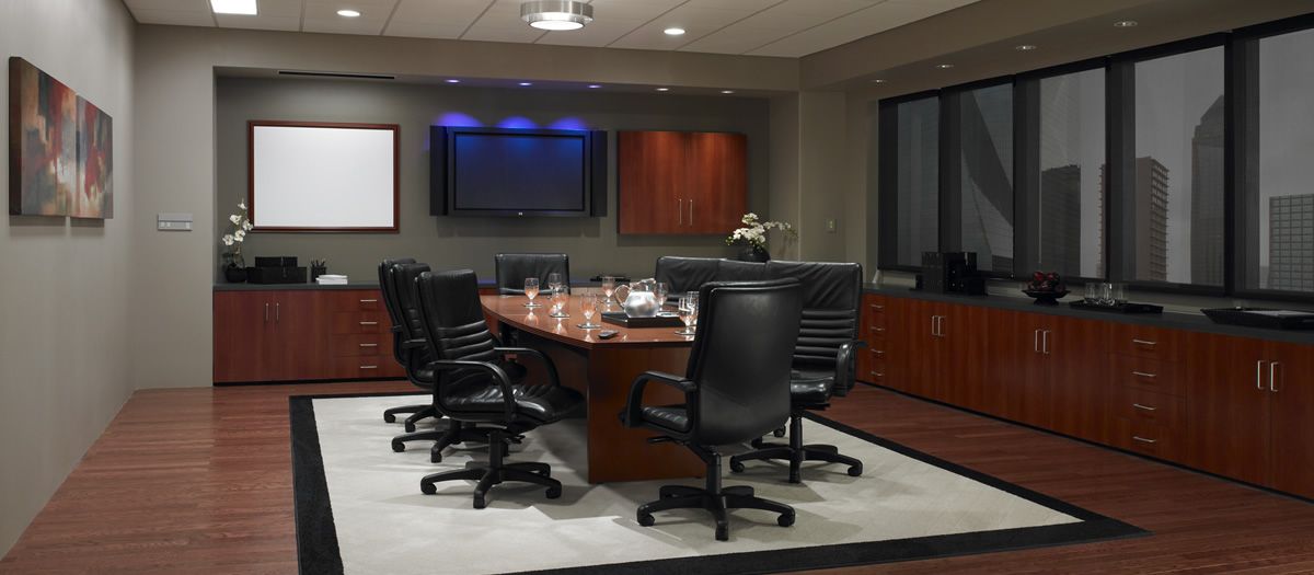 Executive conference room with an oval wood table surrounded by black leather chairs, wall-mounted display and whiteboard, long credenza cabinetry, recessed ceiling lights, and large windows with dark roller shades overlooking a city skyline.